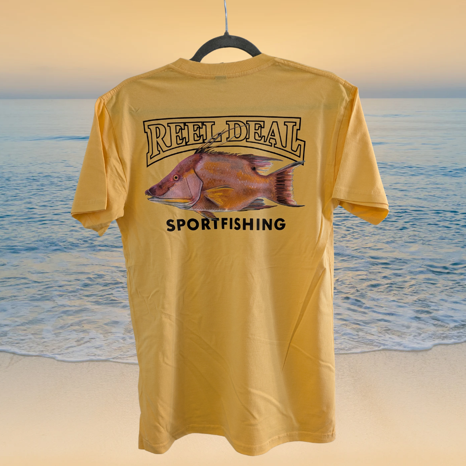 Yellow t-shirt with 'Reel Deal Sportfishing' design on a hanger against a beach background